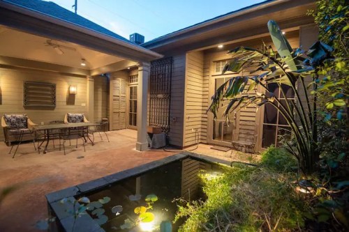 Cozy outdoor patio with seating, lush plants, and a small pond, illuminated in the evening light.