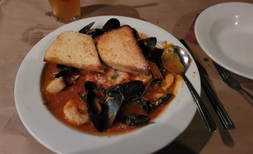 A plate of seafood stew with mussels, shrimp, and toasted bread, served with a spoon on a table.