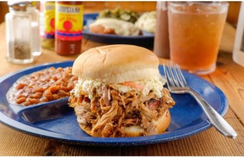 A pulled pork sandwich with coleslaw on a bun, served with baked beans and a drink on a blue plate.
