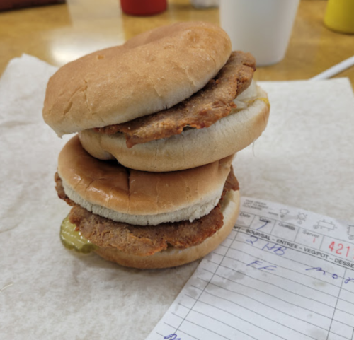 A stacked sandwich with two bread buns and two fried patties, garnished with pickles, on a paper napkin.