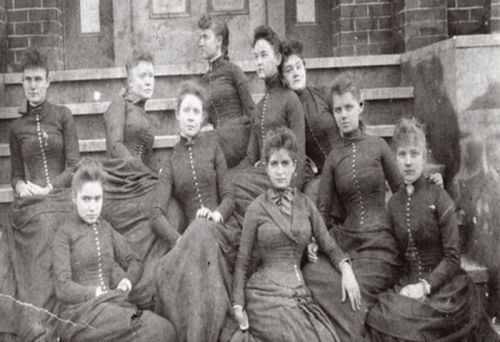 A group of ten women in vintage dresses poses on steps, showcasing 19th-century fashion and hairstyles.