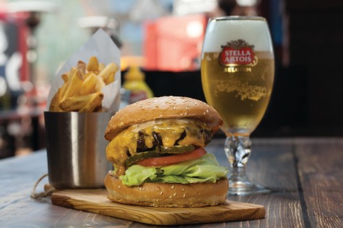 A juicy cheeseburger with lettuce, tomato, and fries, alongside a glass of Stella Artois beer on a wooden table.
