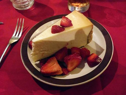 A slice of cheesecake topped with fresh strawberries on a plate, set on a red tablecloth.