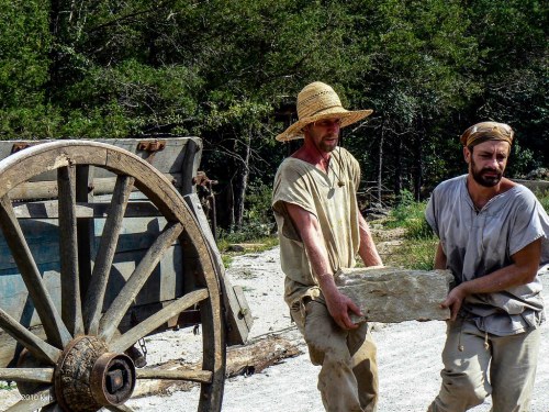 Two men in casual clothing carry a large stone near a wooden cart in a forested area.