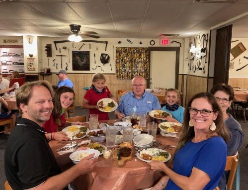 A family gathers around a table, smiling and holding plates of food in a cozy restaurant setting.