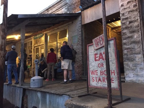 A rustic storefront with people gathered outside, featuring an "Open" sign and a humorous "Eat or We Both Starve" sign.