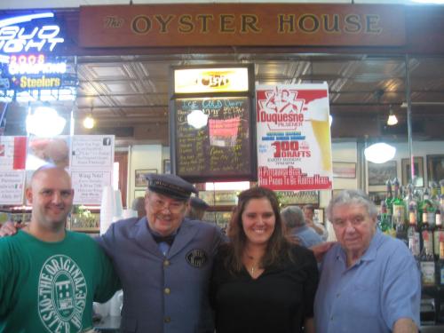A group of four people, including a man in a uniform, poses inside The Oyster House restaurant with a menu board in the background.