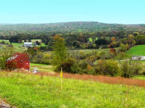 A scenic view of rolling hills with colorful trees, a red barn, and a distant white house under a clear blue sky.