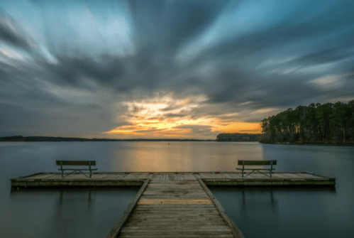 A serene lake at sunset, featuring a wooden dock with two benches and dramatic clouds in the sky.