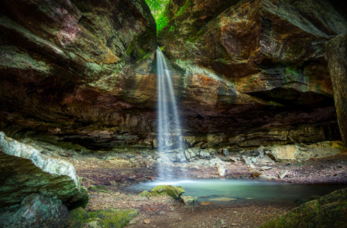 A serene cave with a waterfall cascading down into a tranquil pool, surrounded by rocky walls and lush greenery above.