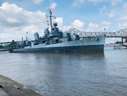 A blue naval ship docked on a river, with a bridge and cloudy sky in the background.