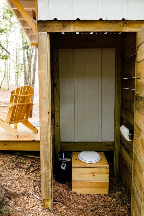 Outdoor restroom with a wooden structure, featuring a composting toilet and an Adirondack chair nearby.