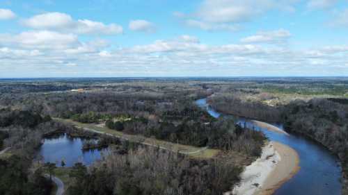 Aerial view of a winding river surrounded by trees and open land under a partly cloudy sky.