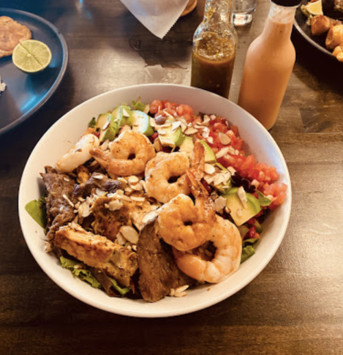 A bowl of salad topped with grilled chicken, shrimp, tomatoes, cucumbers, and sliced almonds, with sauces on the side.