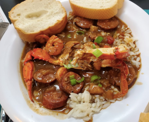 A bowl of gumbo with shrimp, sausage, and crab served over rice, accompanied by two slices of bread.