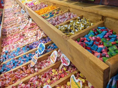 A colorful display of various candies in wooden bins, featuring bright wrappers and playful labels.