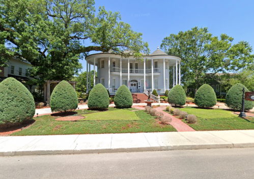 A large, elegant white house with a circular porch, surrounded by neatly trimmed bushes and trees on a sunny day.