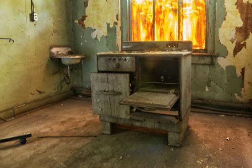 An old, rusty stove stands in a dilapidated room with peeling walls and a window glowing with orange light.