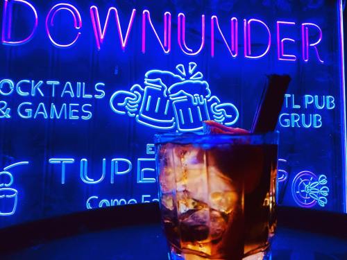A cocktail glass sits in front of a vibrant neon sign that reads "Downunder" with images of beer mugs.