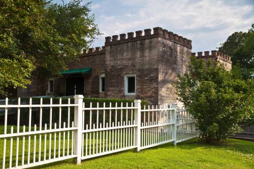 Historic brick building with castle-like features, surrounded by a white picket fence and lush green grass.