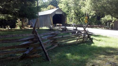 A rustic wooden fence in front of a covered bridge surrounded by trees and greenery on a sunny day.