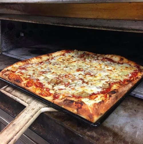 A large rectangular pizza with melted cheese and toppings, partially pulled from a hot oven on a wooden pizza peel.