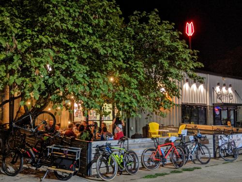 A lively outdoor dining area at night, with bicycles parked under a tree and warm lights illuminating the scene.