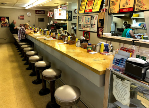 A retro diner interior with a long counter, stools, and condiments, featuring patrons and a staff member in the kitchen.