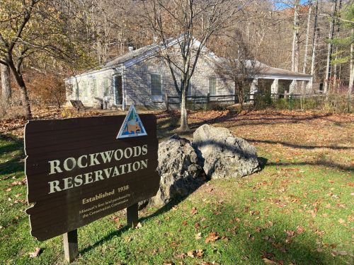 A wooden sign for Rockwoods Reservation in front of a house surrounded by trees and rocks in a natural setting.