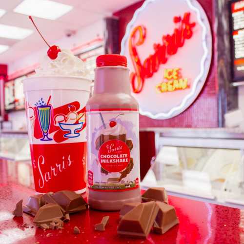 A chocolate milkshake bottle and a cup of milkshake topped with a cherry, set against a bright ice cream shop backdrop.