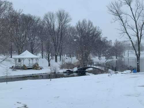 A snowy park scene featuring a gazebo, a bridge over a pond, and bare trees in a winter landscape.