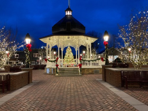 A festive gazebo adorned with lights and a Christmas tree, surrounded by decorated trees and benches at dusk.