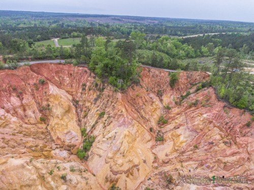 Aerial view of colorful, layered rock formations surrounded by greenery and a winding road in the background.