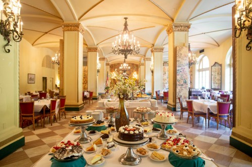 A beautifully arranged dessert table in an elegant dining room, featuring various cakes and pastries under chandeliers.