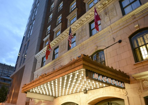 The Magnolia Hotel entrance, featuring bright marquee lights and flags, set against a twilight sky.