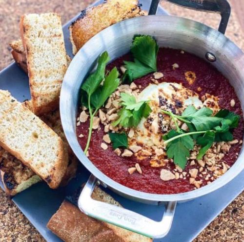 A skillet with baked eggs in a red sauce, topped with herbs and nuts, served with toasted bread on the side.