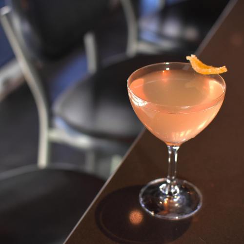 A cocktail in a glass with a twist of orange peel, sitting on a dark bar counter.