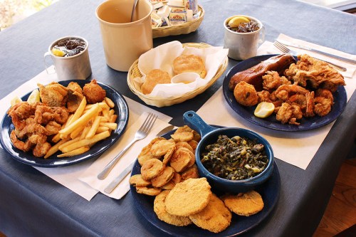 A table set with plates of fried seafood, fries, hushpuppies, collard greens, and drinks.
