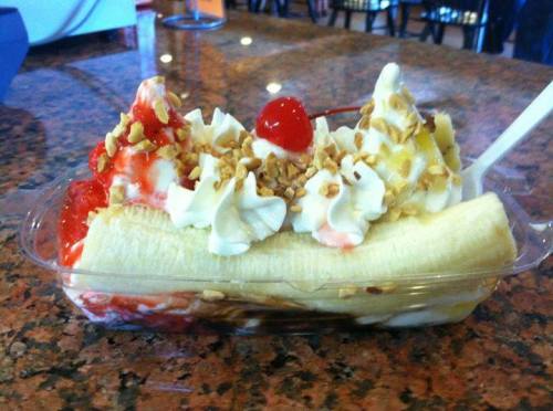 A banana split topped with whipped cream, strawberries, nuts, and a cherry, served in a clear dish on a granite surface.
