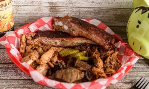 A red and white checkered plate with ribs, pulled pork, and pickles, alongside a drink and a decorative item.