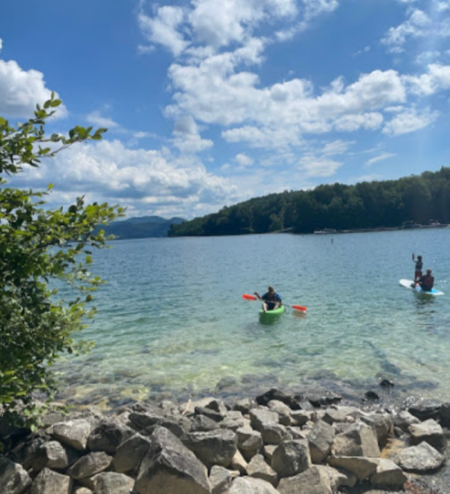 Two people kayaking on a clear lake surrounded by trees and rocky shoreline under a blue sky with clouds.
