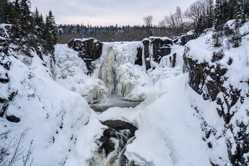 A frozen waterfall surrounded by snow-covered cliffs and trees, creating a serene winter landscape.