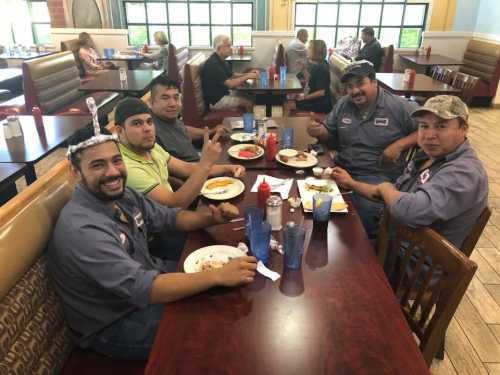 Five men in work uniforms sit at a restaurant table, enjoying meals and drinks, with other diners in the background.