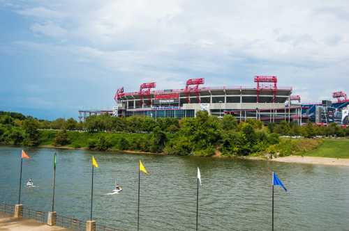 View of a stadium by a river, with colorful flags in the foreground and greenery surrounding the area.