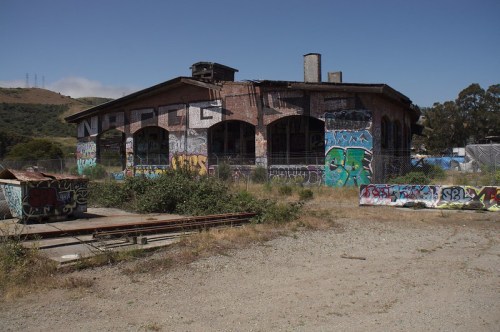 Abandoned building covered in graffiti, surrounded by overgrown grass and debris, with hills in the background.