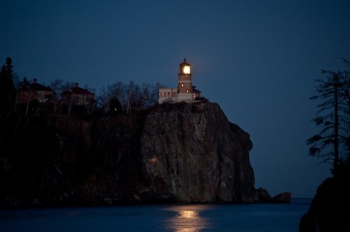 A lighthouse on a rocky cliff illuminated at night, surrounded by trees and reflecting on calm water.