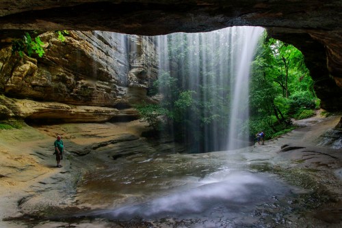A serene waterfall cascades through a rocky overhang, surrounded by lush greenery and a tranquil pool below.
