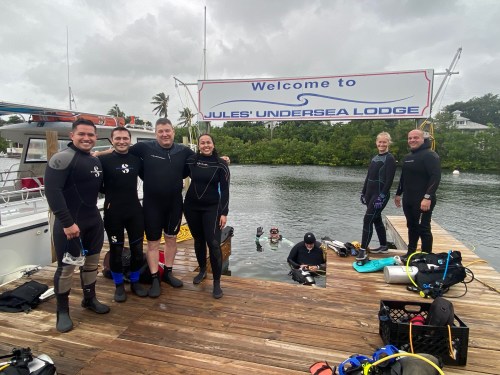 A group of six divers in wetsuits stands on a dock by the water, with a sign reading "Welcome to Jules' Undersea Lodge."