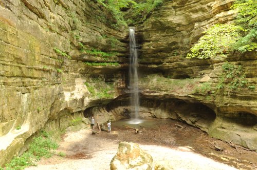 A serene waterfall cascades into a rocky alcove, with two people exploring the area below in a lush, green setting.