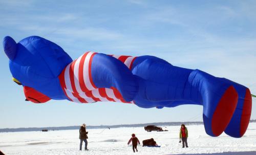 A large blue and red striped balloon character floats above a snowy landscape, with people walking nearby.